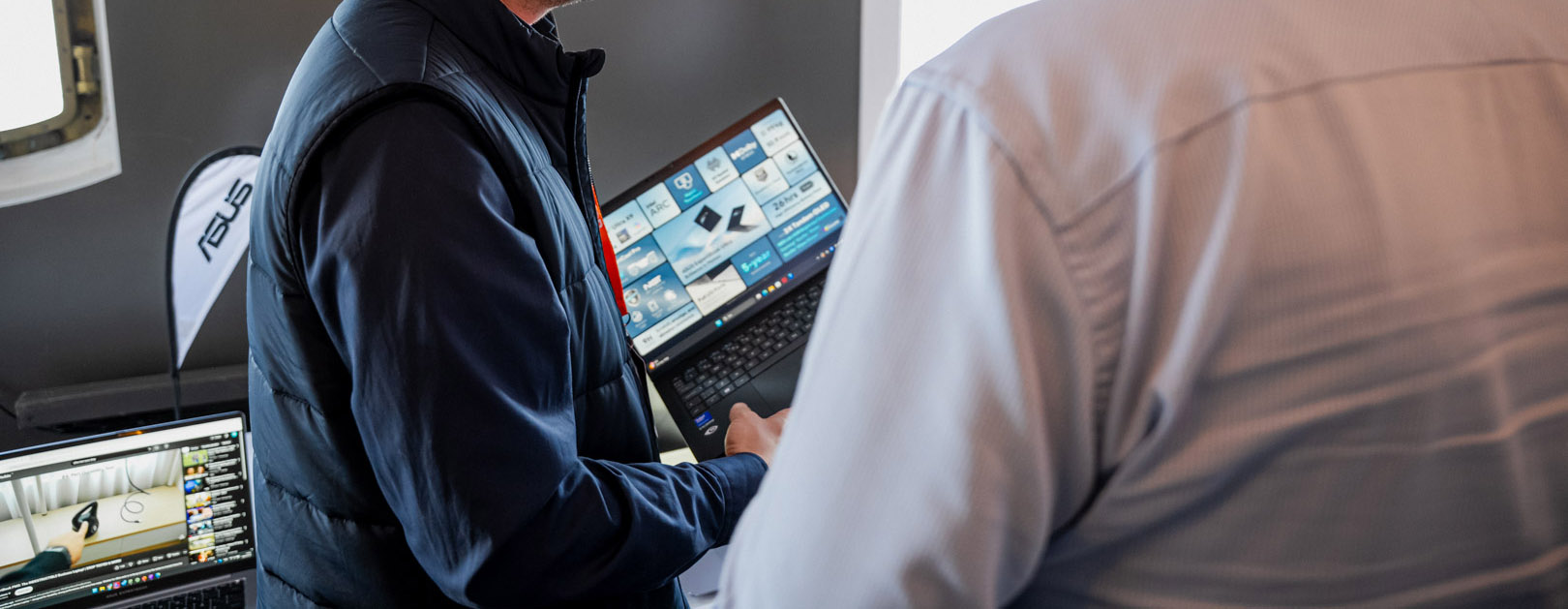 Två personer tittar på en bärbar dator med informationsskärm, ASUS-flagga syns i bakgrunden.
