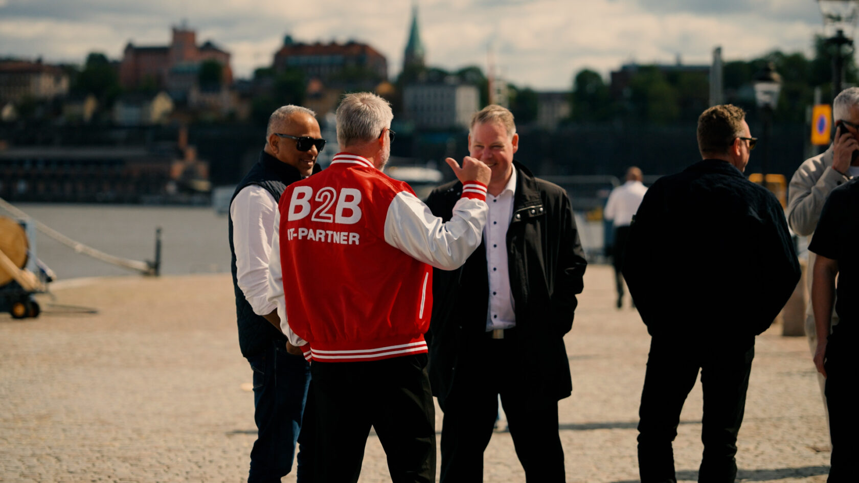 Flera män står och samtalar utomhus vid vattnet, en bär en röd jacka med texten "B2B IT-partner".